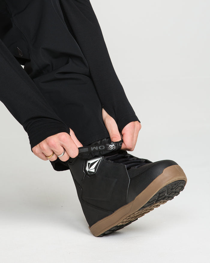 A person wearing black pants styled with the Volcom Men’s V.Co Sparta Bib fastens the strap on a black boot with a brown sole and white logo, against a plain white background.