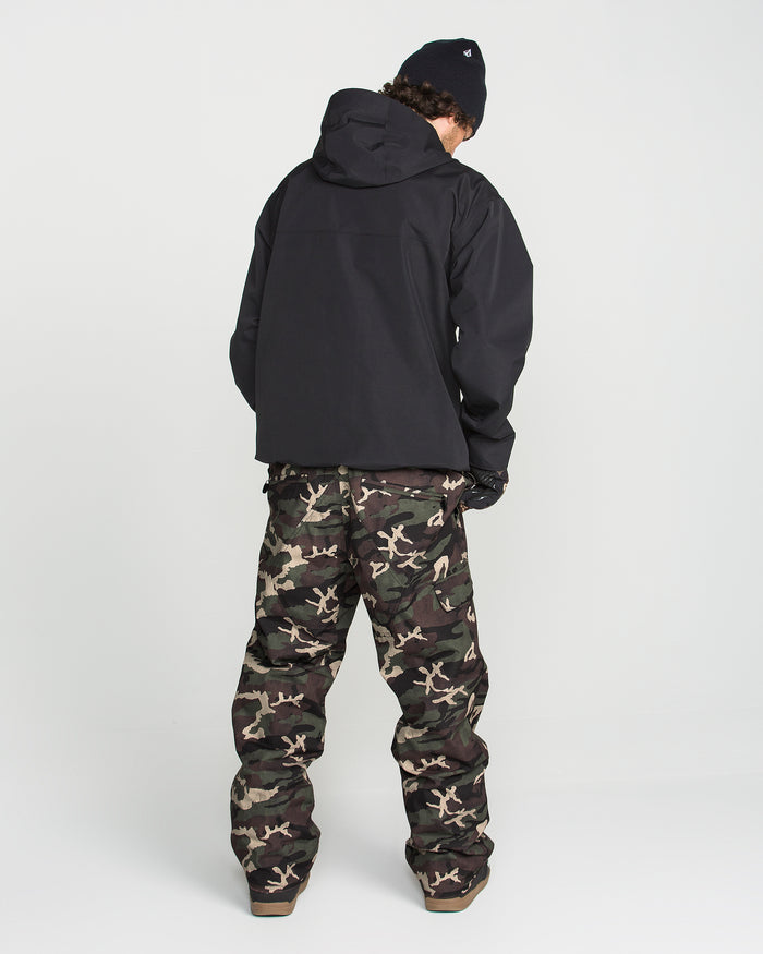 A person stands with their back to the camera, wearing the Volcom Men’s V.Co Sparta Bib, paired with a black hooded jacket made from recycled fabric, a black knit hat, and gloves against a plain white background.