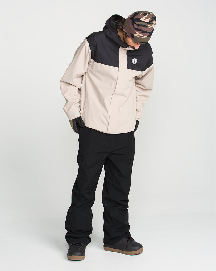 A person wearing Volcom’s Men’s V.Co Sparta Bib in tan and black recycled fabric, paired with black pants, black gloves, a black-and-tan camo beanie, and dark sneakers stands looking down against a white background.