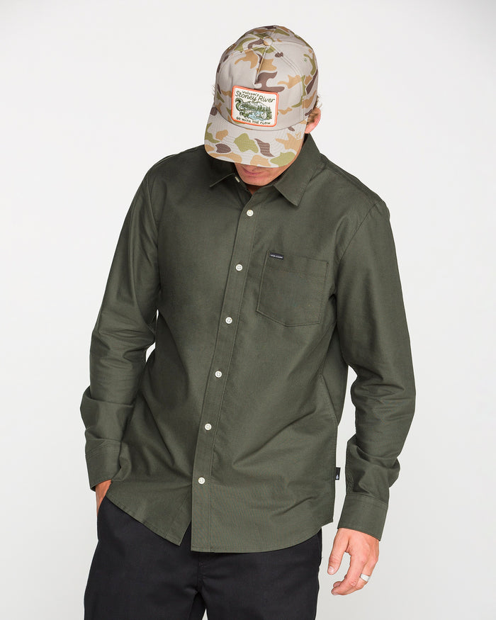 A person wearing the Volcom Men's Veeco Oxford Long Sleeve Shirt in Climbing Ivy green, paired with black pants and a camouflage baseball cap with a label, looks down with one hand in a pocket against a plain light background.