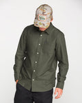 A person wearing the Volcom Men's Veeco Oxford Long Sleeve Shirt in Climbing Ivy green, paired with black pants and a camouflage baseball cap with a label, looks down with one hand in a pocket against a plain light background.