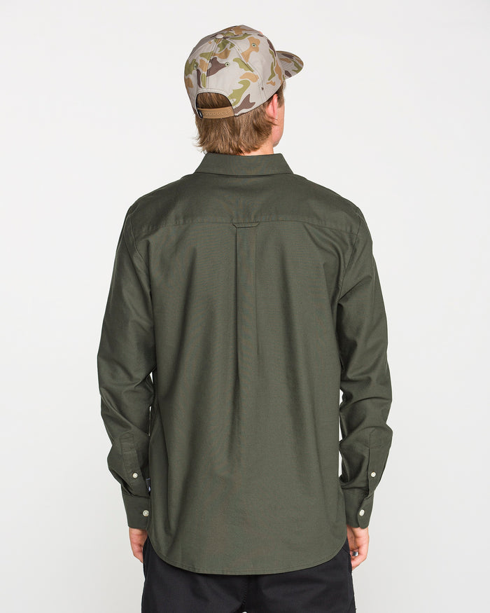 A person wearing a Climbing Ivy green Men's Veeco Oxford Long Sleeve Shirt in a classic fit by Volcom and a camouflage baseball cap stands facing away, displaying the back of their outfit against a plain white background.