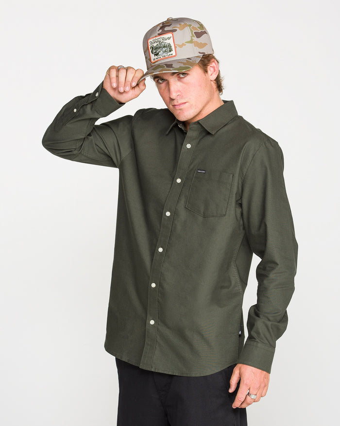 A person wearing the Volcom Men's Veeco Oxford Long Sleeve Shirt in Climbing Ivy green and black pants adjusts a camouflage baseball cap. The classic fit and micro suede fabric of the shirt add a subtle touch of sophistication against the plain white background.