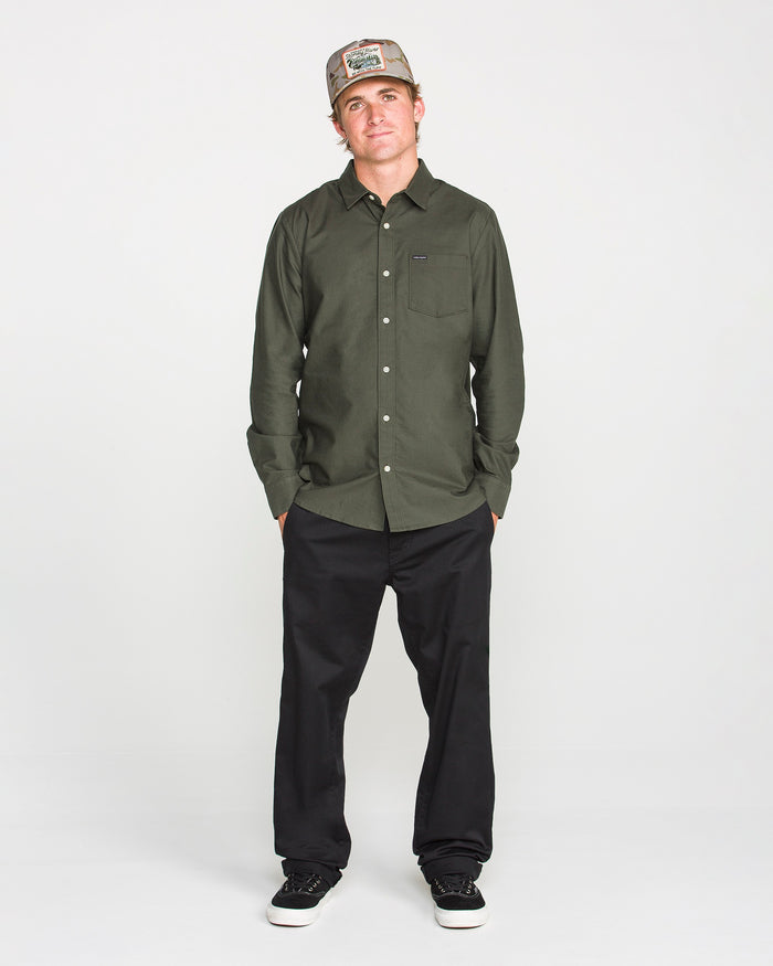A man in a Volcom Men's Veeco Oxford Long Sleeve Shirt in Climbing Ivy green, classic fit black pants, and black sneakers stands with his hands in his pockets, wearing a tan baseball cap against a plain white background.
