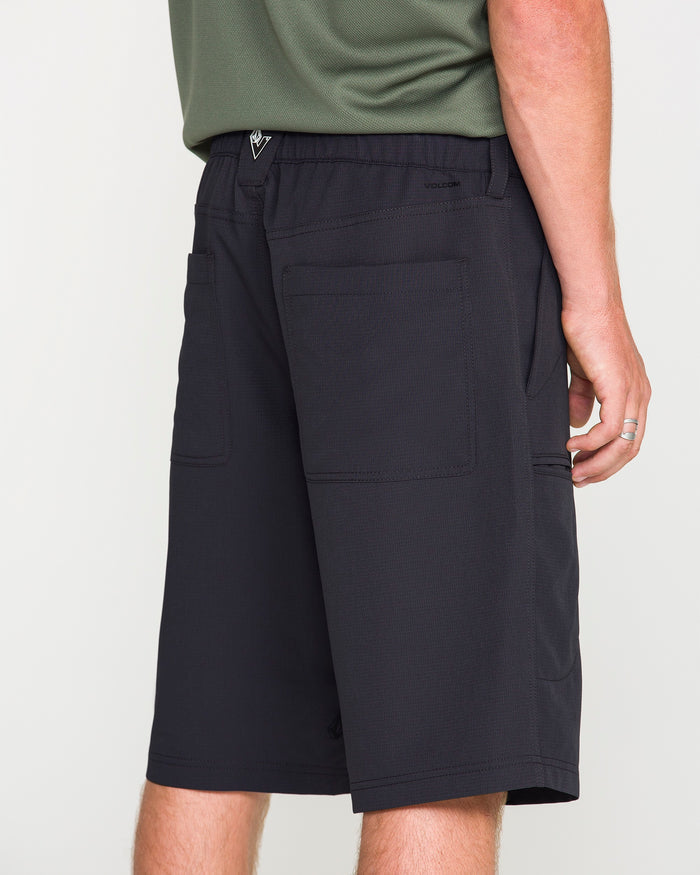 A person wearing Volcom Men's Venture Stone Hybrid Shorts in Asphalt Black, featuring knee-length recycled-poly fabric and large back pockets, along with a green shirt and a silver ring, standing against a plain white background.