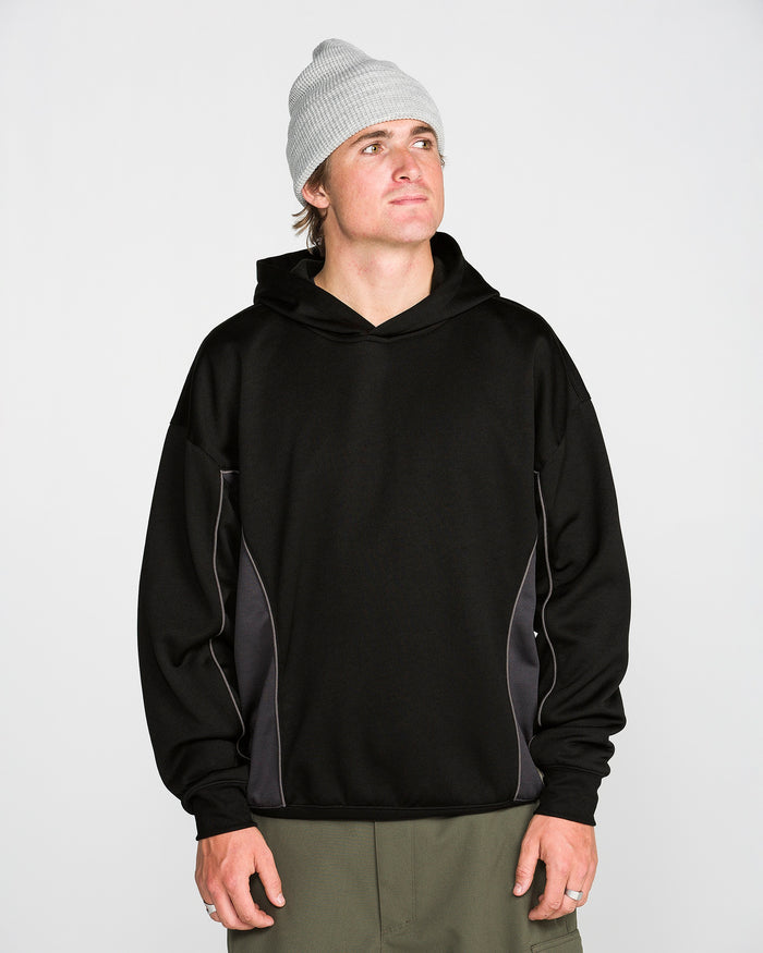 A person wearing a light gray beanie, olive green pants, and the Volcom Men’s Vital Hydro Pullover Hoodie in black with gray side panels stands against a plain white background, looking slightly upward and to the right.
