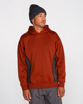 A man wearing the Volcom Men’s Vital Hydro Pullover in rust—with contrasting black side panels—pairs it with gray pants and a dark knit beanie as he stands against a plain light background, gazing to his left. The cozy, hydrophobic fleece of the pullover delivers both style and comfort, reflecting Volcom’s modern appeal.