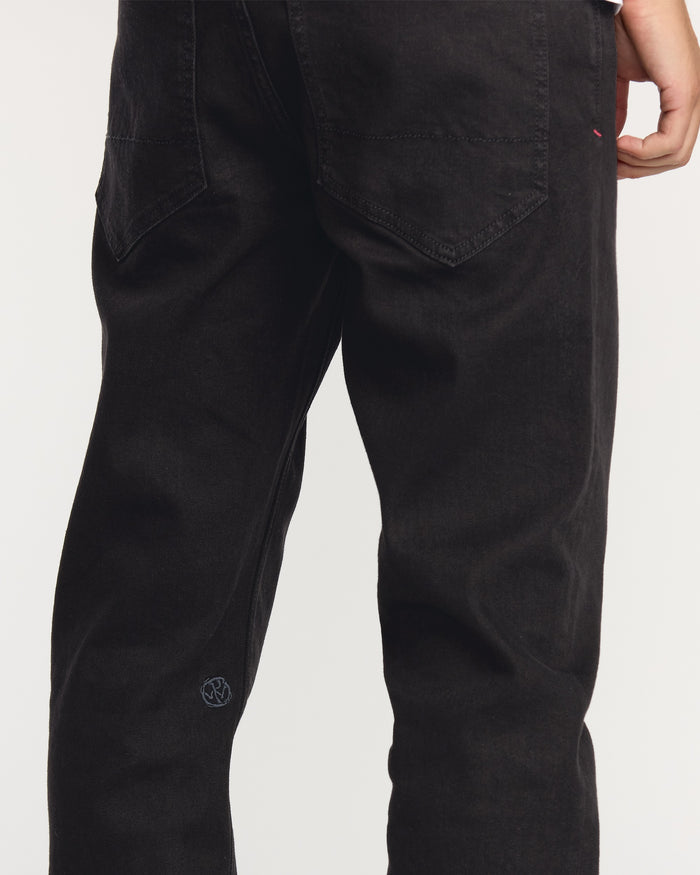 A person is shown from the back wearing Volcom Men's Volcom Entertainment Pennywise Denim Pants. The black jeans feature visible stitching and back pockets, with one hand resting at their side against a plain light background.