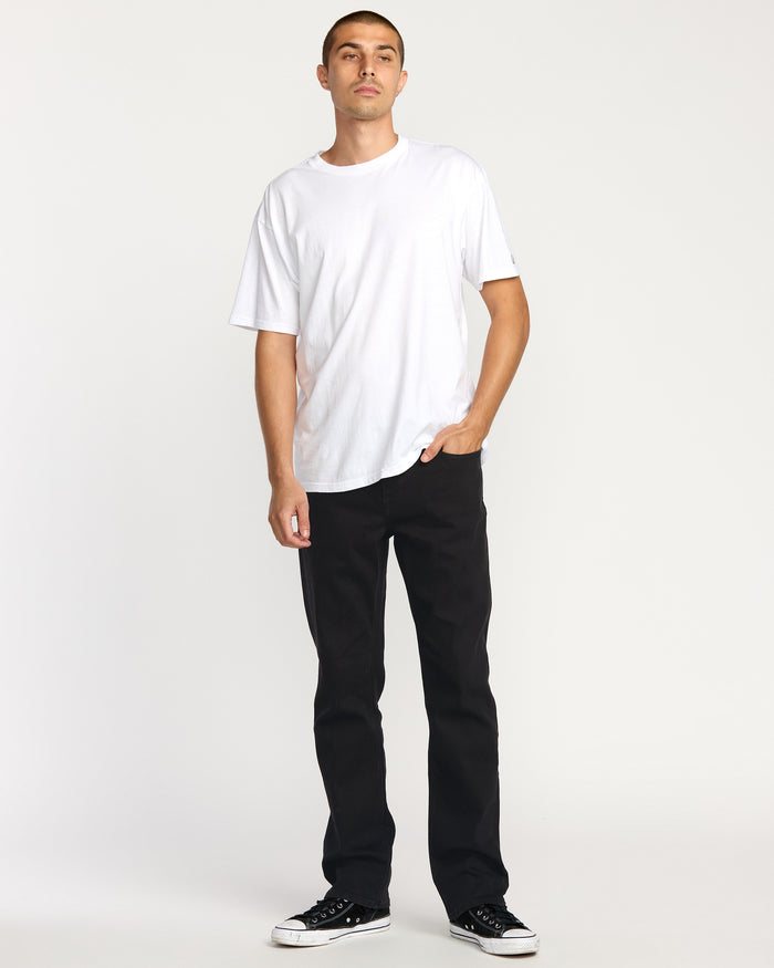 A man stands against a white background, wearing Volcom's Men's Volcom Entertainment Pennywise Denim Pants with a white short-sleeve t-shirt and black sneakers. One hand is in his pocket, and he has a neutral expression.