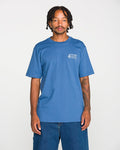 A young Black man with braided hair and a mustache stands against a white background, wearing the Volcom Men's Volcom Workwear Short Sleeve Tee in blue paired with blue jeans. He has visible tattoos on his left arm and is looking directly at the camera.
