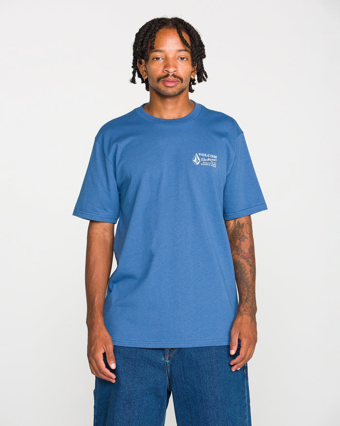 A young Black man with braided hair and a mustache stands against a white background, wearing the Volcom Men's Volcom Workwear Short Sleeve Tee in blue paired with blue jeans. He has visible tattoos on his left arm and is looking directly at the camera.