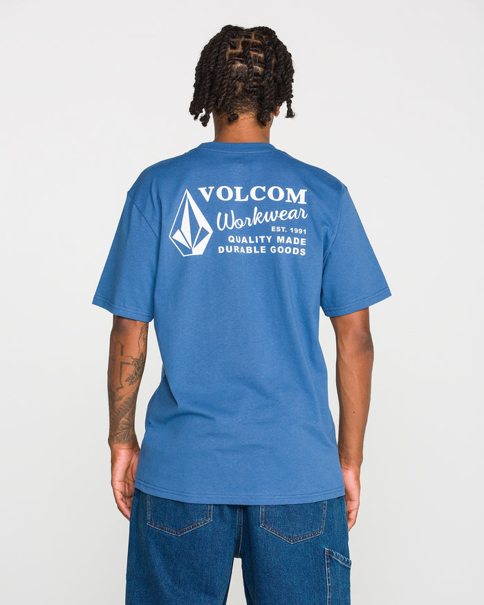 A person is seen from behind wearing blue jeans and the Men’s Volcom Workwear Short Sleeve Tee by Volcom, a classic fit in blue crafted from heavyweight cotton. The back of the shirt displays a white Volcom logo with the text “Quality Made Durable Goods.”.
