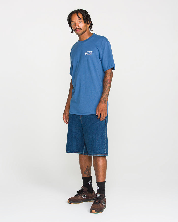 A man with braided hair wears the Men's Volcom Workwear Short Sleeve Tee in blue, styled with long denim shorts, black socks, and brown sneakers as he stands against a plain white background—a classic Volcom look.