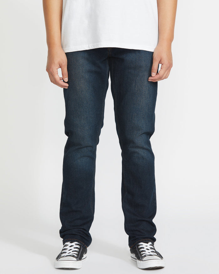 Wearing Volcom's Men's Vorta Denim jeans, a person stands against a plain white background. The image is cropped from the shoulders down and also shows a white t-shirt and black-and-white sneakers.