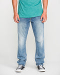 A person wearing a green t-shirt, Volcom Men's Vorta Denim Pants in a slim fit, and black-and-white sneakers stands with arms relaxed at their sides against a plain background.