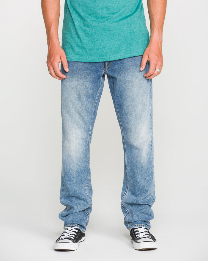 A person wearing a green t-shirt, Volcom Men's Vorta Denim Pants in a slim fit, and black-and-white sneakers stands with arms relaxed at their sides against a plain background.