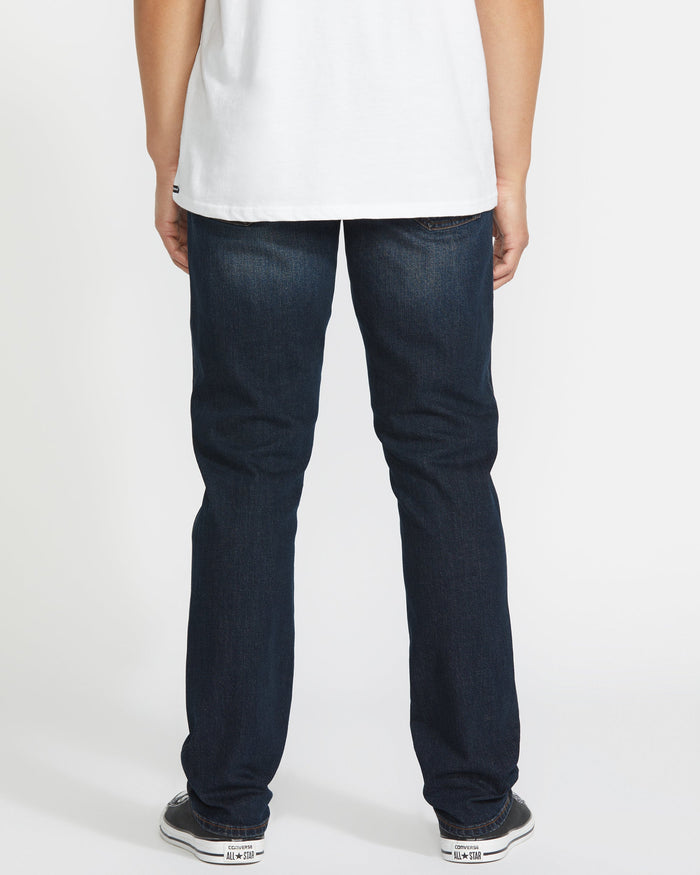 A person stands with their back to the camera, wearing a white t-shirt, Volcom Men's Vorta Denim dark blue jeans, and black and white sneakers against a plain white background.