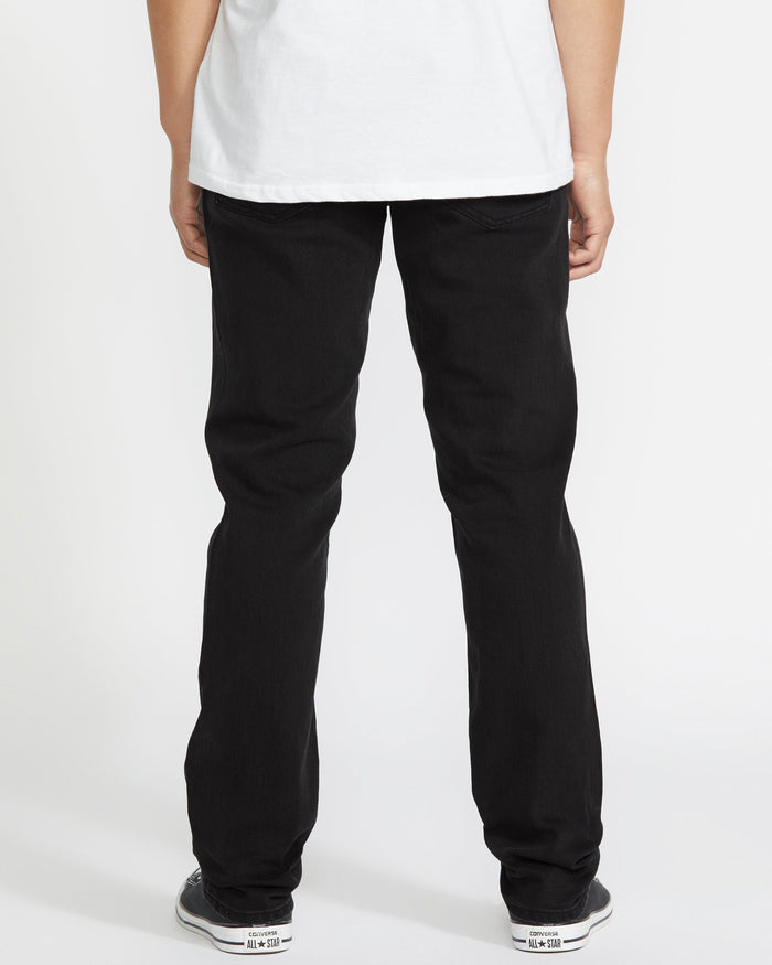 A person faces away from the camera, wearing a white shirt, Volcom Vorta Denim black stretch straight leg jeans, and black-and-white sneakers against a plain white background.