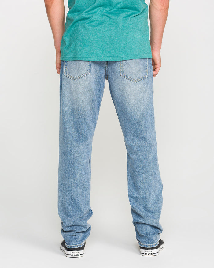 A person wearing a green T-shirt, Volcom Men's Vorta Denim Pants in Freedom Blue (slim fit), and black-and-white sneakers stands facing away, showcasing the back view of the jeans.