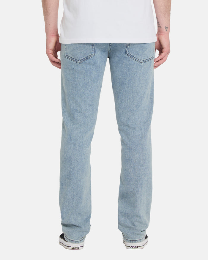 A person wearing Volcom Men's Vorta Denim Pants in light blue, a white T-shirt, and black-and-white sneakers stands with their back to the camera against a plain white background.