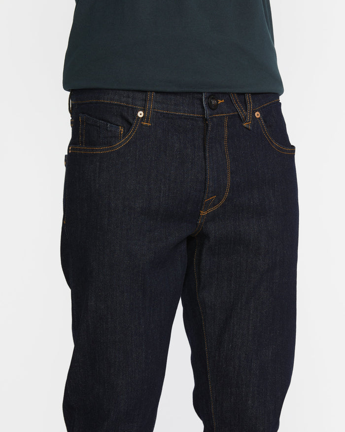A person wears slim fit Men's Vorta Denim by Volcom with orange stitching and a dark green shirt. The photo focuses on the waist and upper legs against a white background, highlighting Water Aware details.