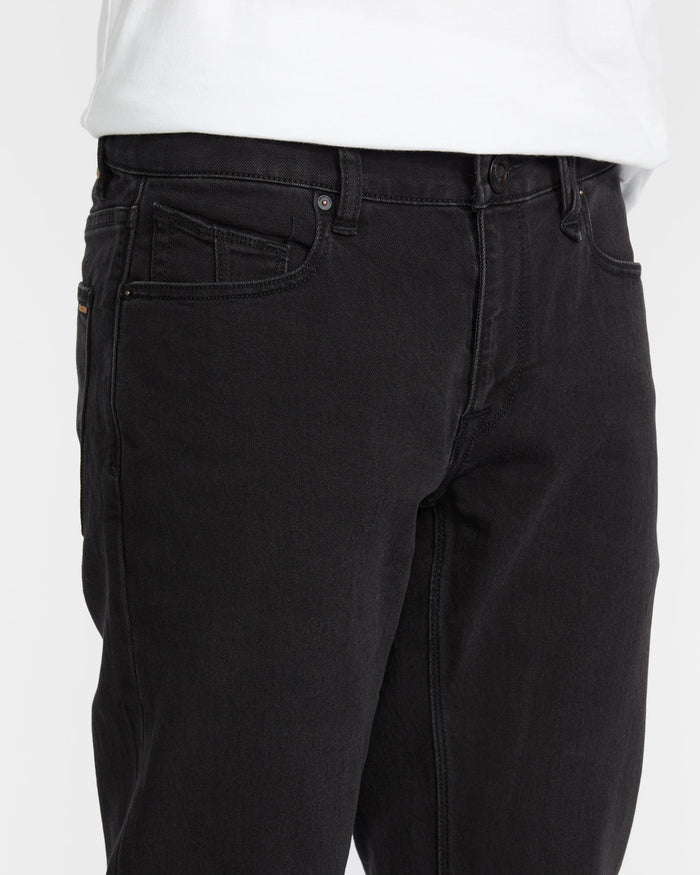 A person wearing Volcom Vorta Denim black slim fit jeans made from stretch denim and a white shirt, shown from waist to mid-thigh with their left hand out of view.