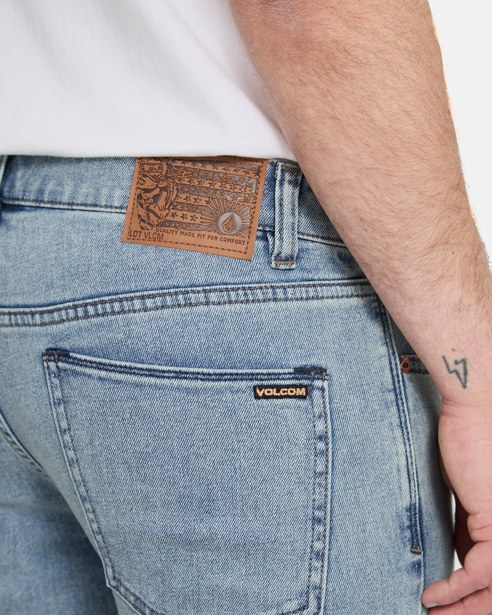 Close-up of a person wearing Volcom Men's Vorta Denim Pants in light blue, with a brown leather patch on the waistband and Volcom label above the back pocket. A white shirt and arm tattoo are partially visible.