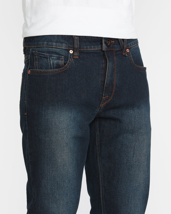 A person wearing Volcom Men's Vorta Denim jeans in dark blue with orange stitching and a white shirt. The image highlights the waist, upper legs, front pockets, and button closure.