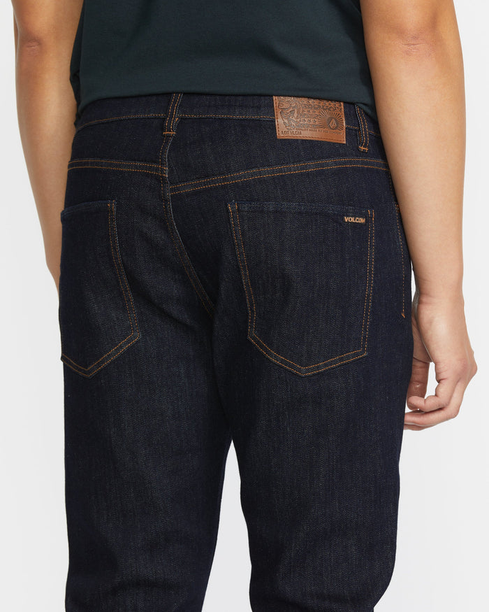 A person is seen from the back wearing Volcom Men's Vorta Denim jeans in dark blue with orange stitching, a brown leather waistband patch, and a Volcom label above the right pocket, paired with a dark green shirt.
