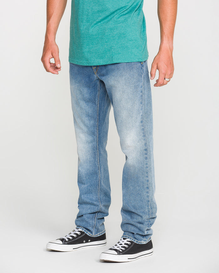 A person wearing a green t-shirt, Volcom Men's Vorta Denim Pants in a slim fit, and black Converse sneakers stands against a plain white background. Their face is not visible.