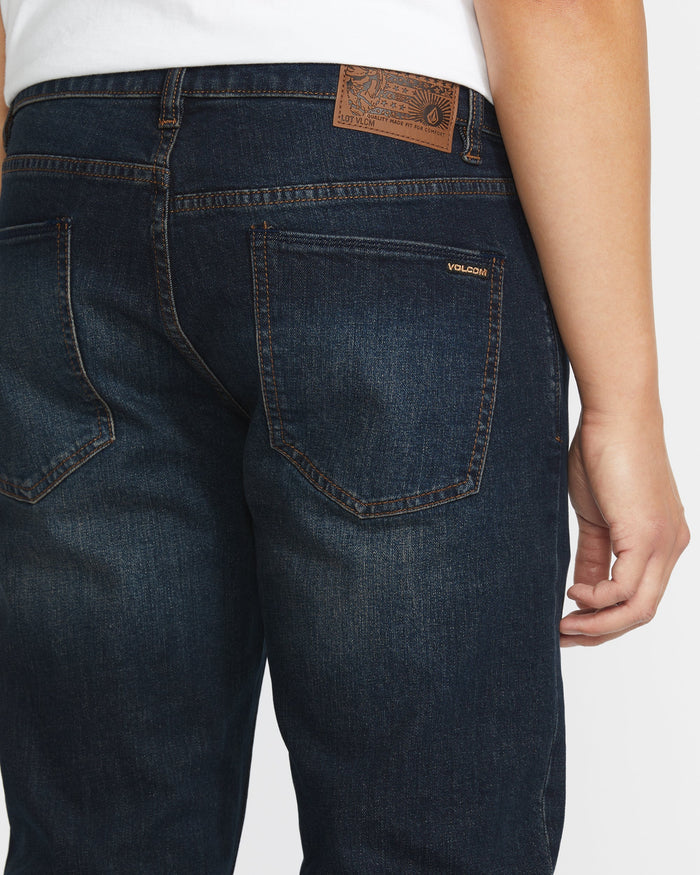 Close-up of the back of a person wearing Volcom Men's Vorta Denim in dark blue with brown stitching, a brown leather patch on the waistband, and Volcom branding above the right back pocket. The person is also wearing a white shirt.