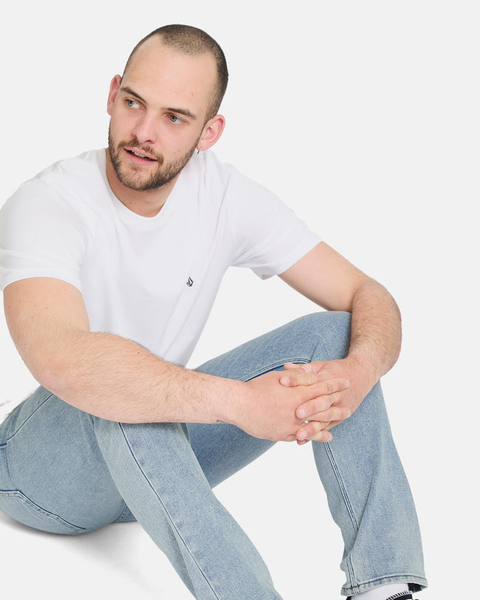 A man with a shaved head and beard sits cross-legged on the floor, wearing a white t-shirt and Volcom Men's Vorta Denim Pants, looking relaxed as he gazes off to the side.