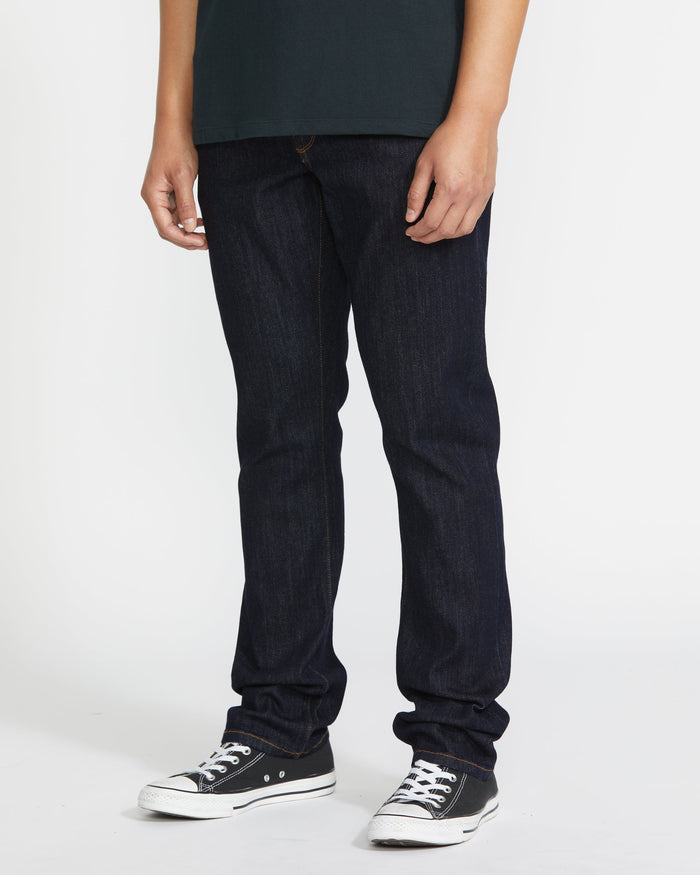 A person wearing a dark green shirt, Volcom Men's Vorta Denim in slim fit, and black low-top sneakers stands against a white background. The cropped image from shoulders down highlights their water aware style.