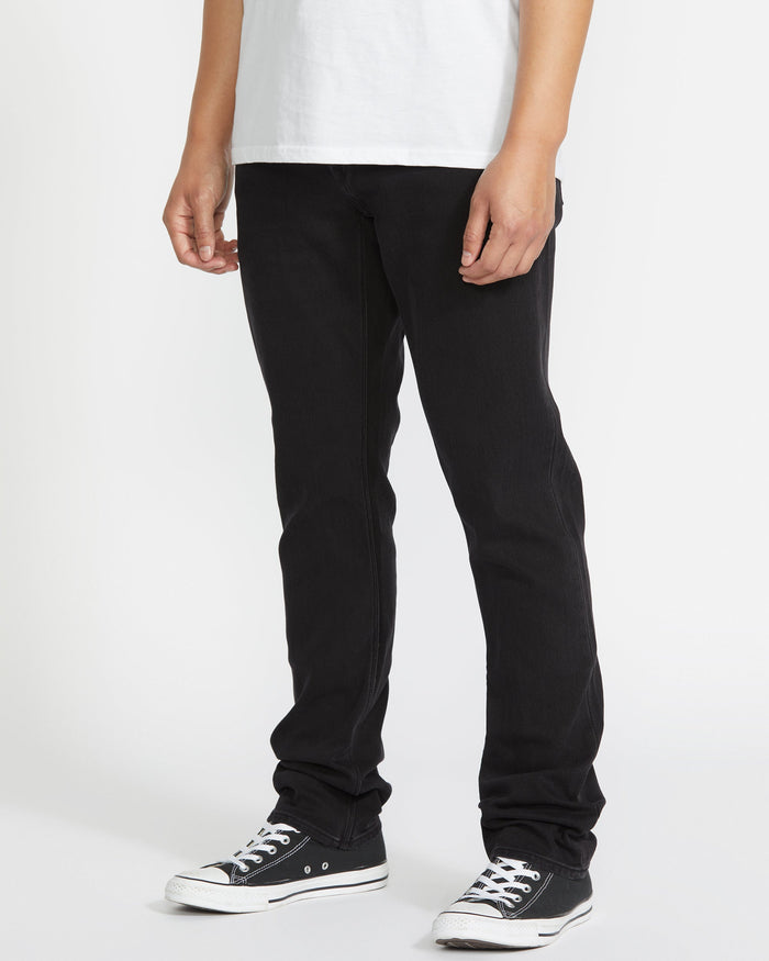 Only the lower half of the body is visible, showing a person standing against a white background wearing a white t-shirt, Volcom Vorta Denim black straight leg stretch jeans, and black low-top sneakers with white soles.