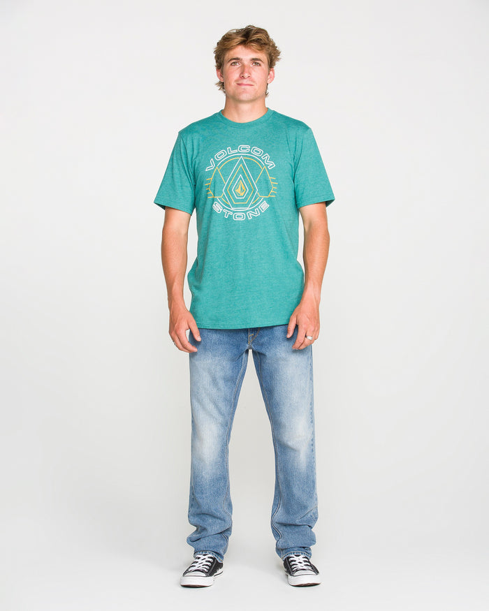 A young man stands against a plain white background, wearing a teal graphic T-shirt, Volcom Men's Vorta Denim Pants in Freedom Blue, and black-and-white sneakers. He is facing forward with his arms relaxed at his sides.