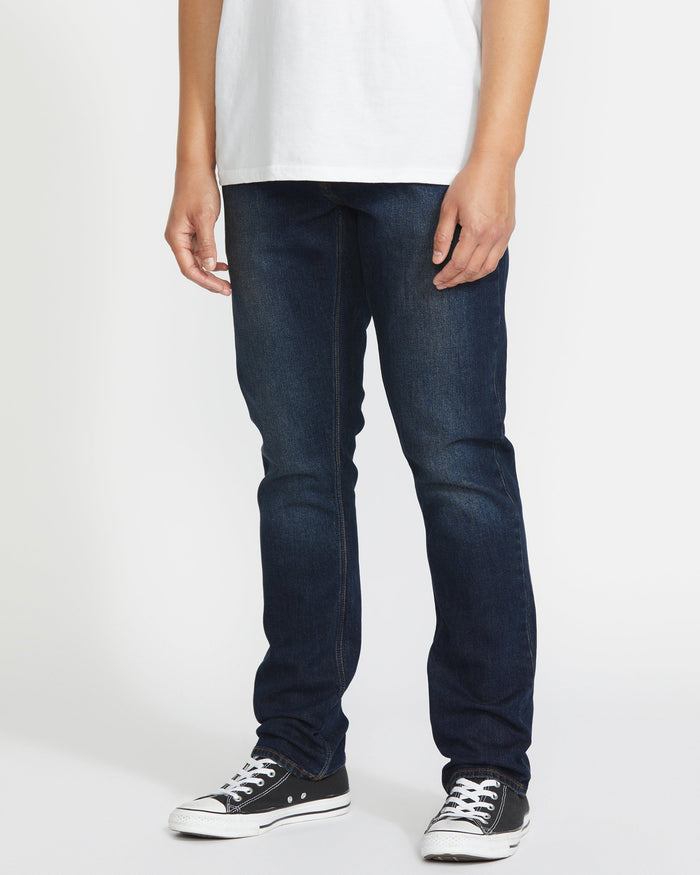A person stands against a plain white background, showing only their lower half while wearing Volcom Men's Vorta Denim jeans, a plain white t-shirt, and black low-top Converse sneakers.