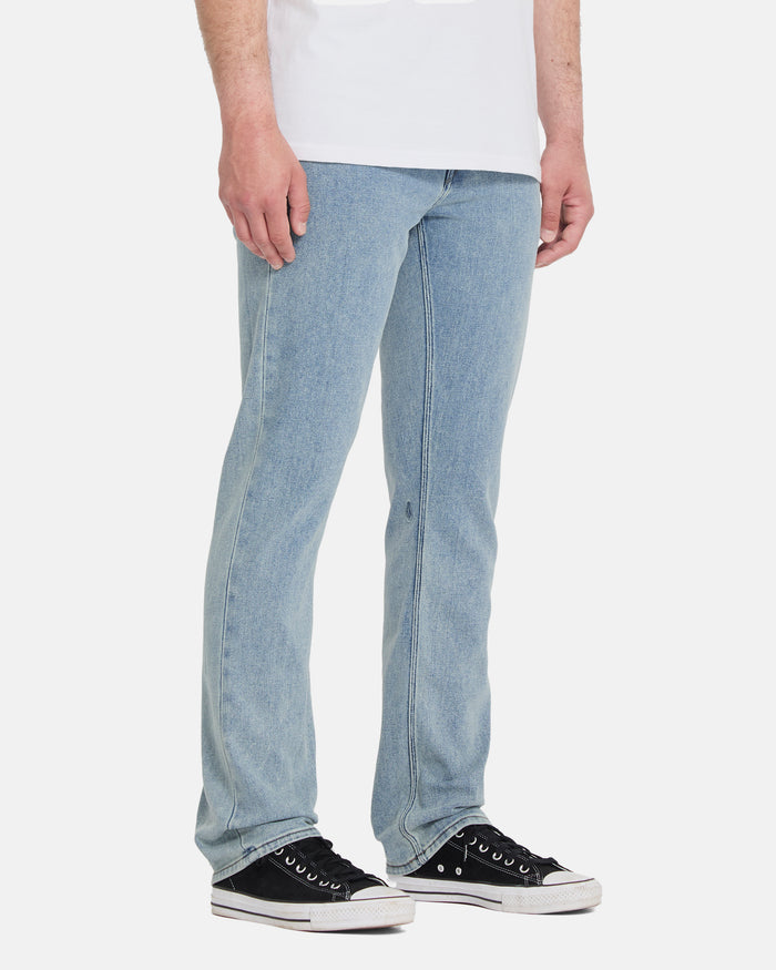A person wearing light blue Volcom Men's Vorta Denim Pants, a white shirt, and black low-top sneakers with white soles stands against a plain white background.