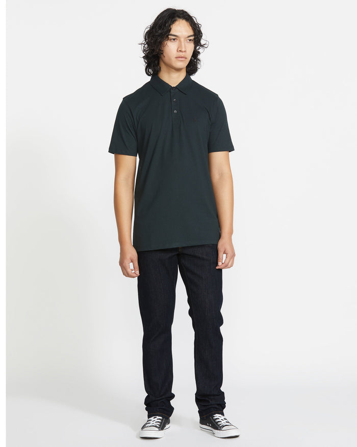 A young person with medium-length curly hair, wearing a dark polo, Volcom Men's Vorta Denim in a slim fit, and black sneakers stands against a plain white background.