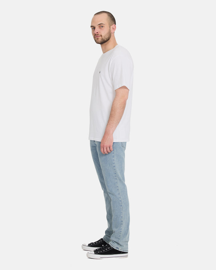 A bearded man in profile wears a white t-shirt, Volcom Men's Vorta Denim Pants in light blue, and black sneakers against a plain white background.