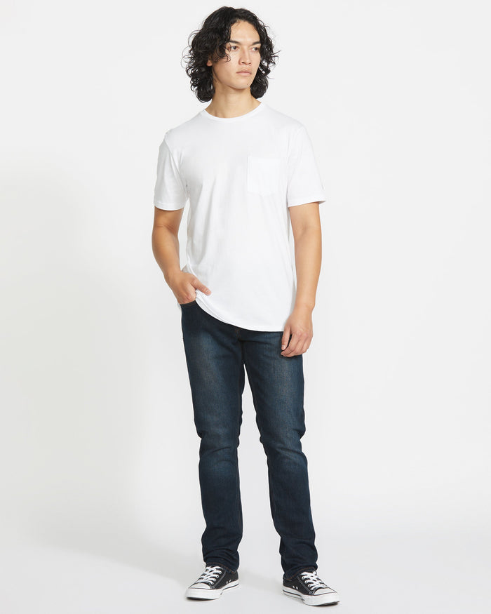 A person with curly hair stands against a white background, wearing a white short-sleeve t-shirt, Volcom Men's Vorta Denim jeans in dark blue, and black-and-white sneakers, with one hand in their pocket.