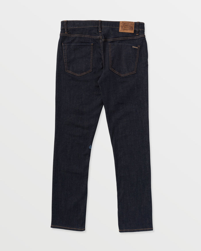 A pair of Men's Vorta Denim jeans by Volcom in dark blue slim fit is shown from the back with two rear pockets, brown stitching, and a tan leather patch on the waistband. The Water Aware jeans are laid flat on a white background.