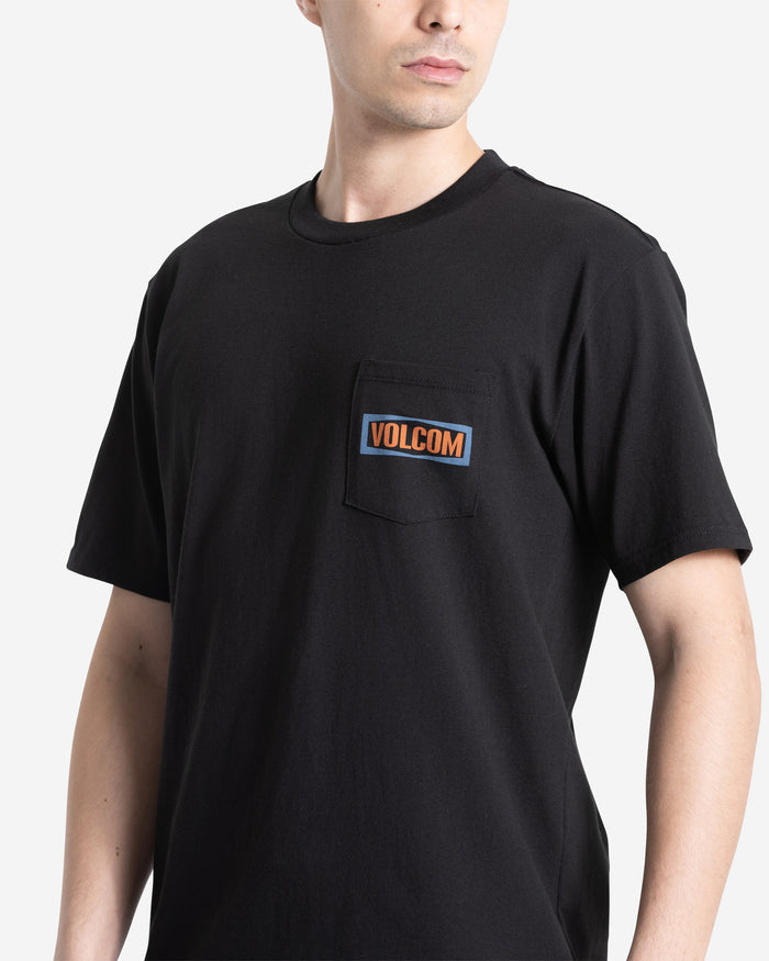 A person wears the Volcom Men's Warning Pocket Short Sleeve Tee, a black 100% cotton shirt with a chest pocket featuring an orange and white VOLCOM patch, shown against a plain light background.