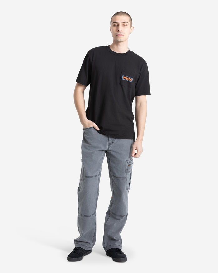 A person stands against a white background wearing a Volcom Men's Warning Pocket Short Sleeve Tee, black with a chest patch, paired with gray cargo pants and black shoes. One hand is in their pocket and they have a neutral expression.