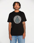 A man with short dreadlocks stands facing the camera against a plain white background, wearing blue jeans and the Volcom Men's Wave Stone Short Sleeve Tee—a black, premium ring spun cotton T-shirt featuring a geometric leaf design.