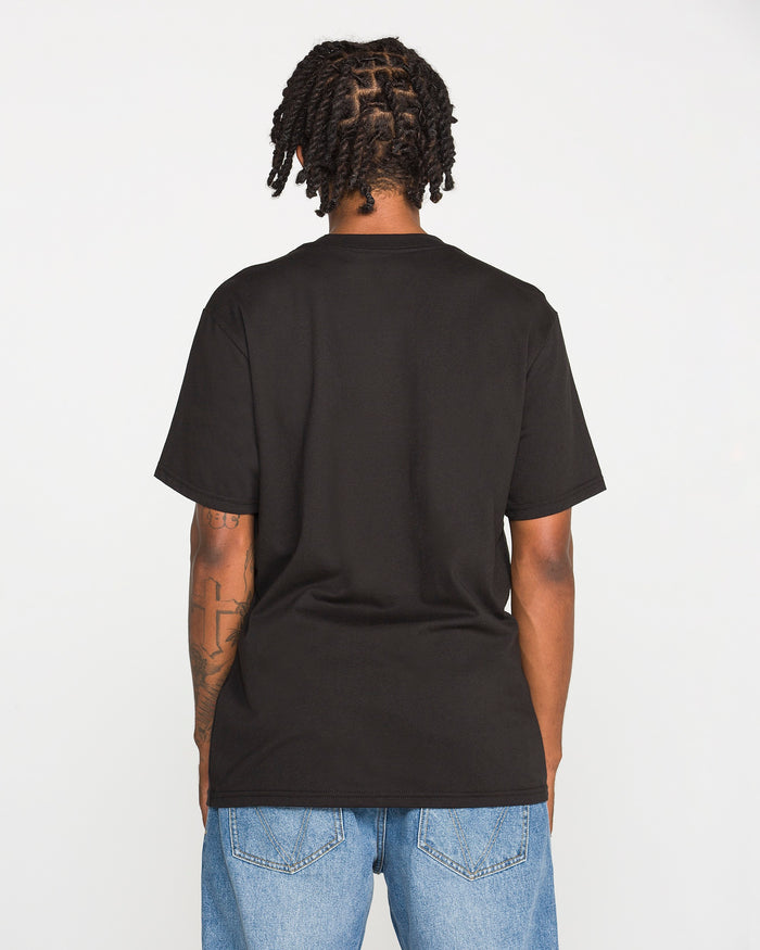 A person with short, twisted hair stands facing away, wearing the Volcom Men's Wave Stone Short Sleeve Tee in plain black, crafted from premium ring-spun cotton in a classic fit, paired with blue jeans. Their arms are visible, showcasing tattoos on the left arm. The background is plain white.