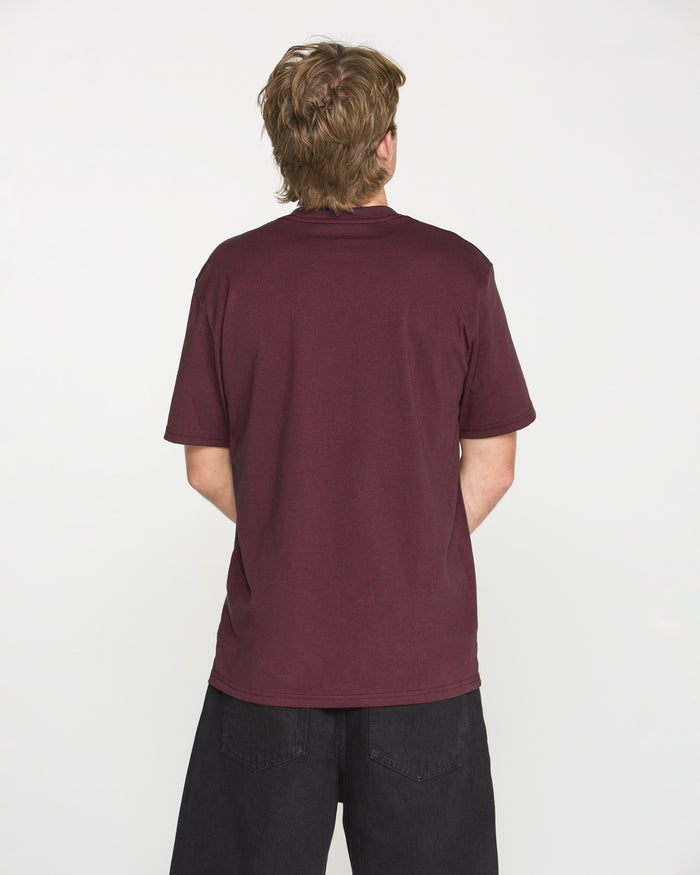 A person with short, light brown hair stands facing away, wearing the Volcom Men's Wave Stone Short Sleeve Tee in Burgundy, crafted from premium ring spun cotton, paired with loose black pants against a plain white background.