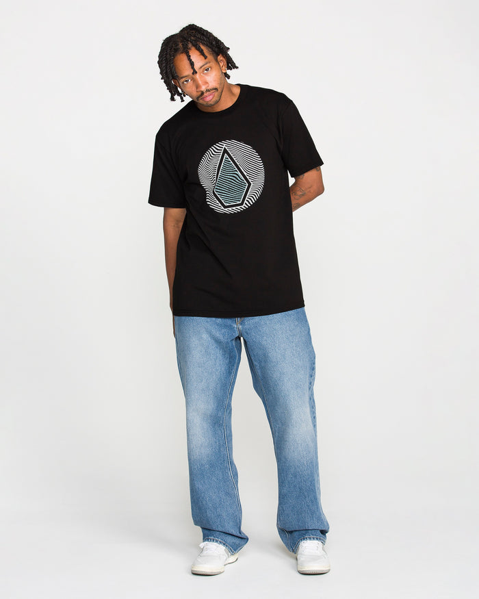 A man with short dreadlocks wears the Volcom Men's Wave Stone Short Sleeve Tee in black, paired with blue jeans and white sneakers. He stands against a plain white background, slightly tilting his head while leaning with one hand behind his back.