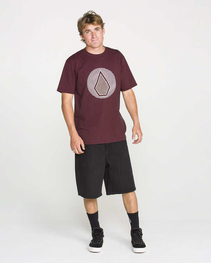 A man stands against a plain white background wearing the Volcom Men's Wave Stone Short Sleeve Tee in Burgundy, crafted from premium ring spun cotton and featuring a geometric logo, paired with black shorts, black socks, and black sneakers.