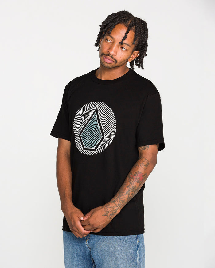 A man with braided hair, a mustache, and tattoos on his forearm wears the Volcom Men's Wave Stone Short Sleeve Tee—a classic fit t-shirt made from premium ring spun cotton and featuring a geometric design. He pairs it with blue jeans and stands against a plain white background.
