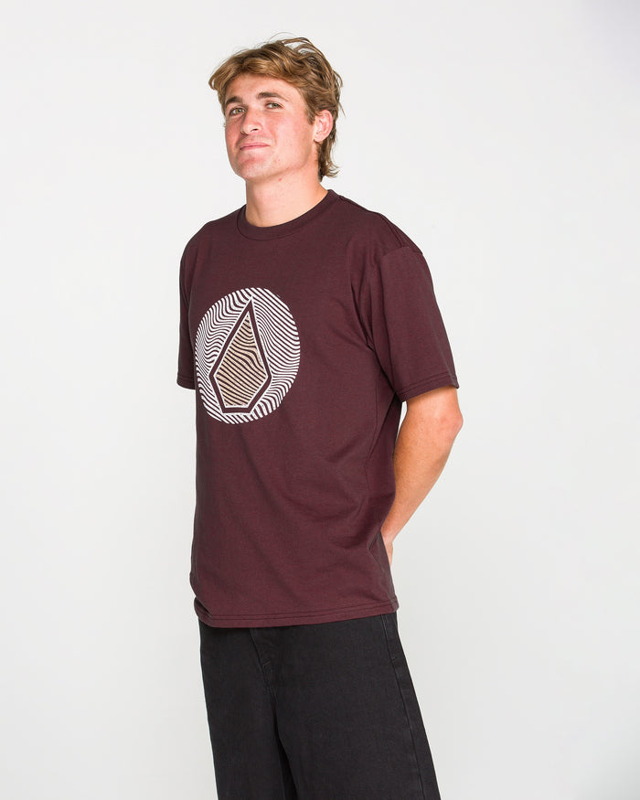 A person with short blond hair wears the Volcom Men's Wave Stone Short Sleeve Tee—a premium ring spun cotton burgundy t-shirt featuring a geometric circular logo—paired with black pants, standing against a plain white background with their hands behind their back.